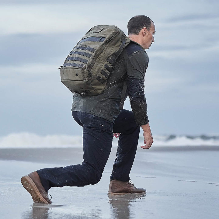 A person kneels on the beach with a green backpack, facing ocean waves beneath a cloudy sky, wearing GORUCK's MACV-1 Hi-Speed Mid Top boots.