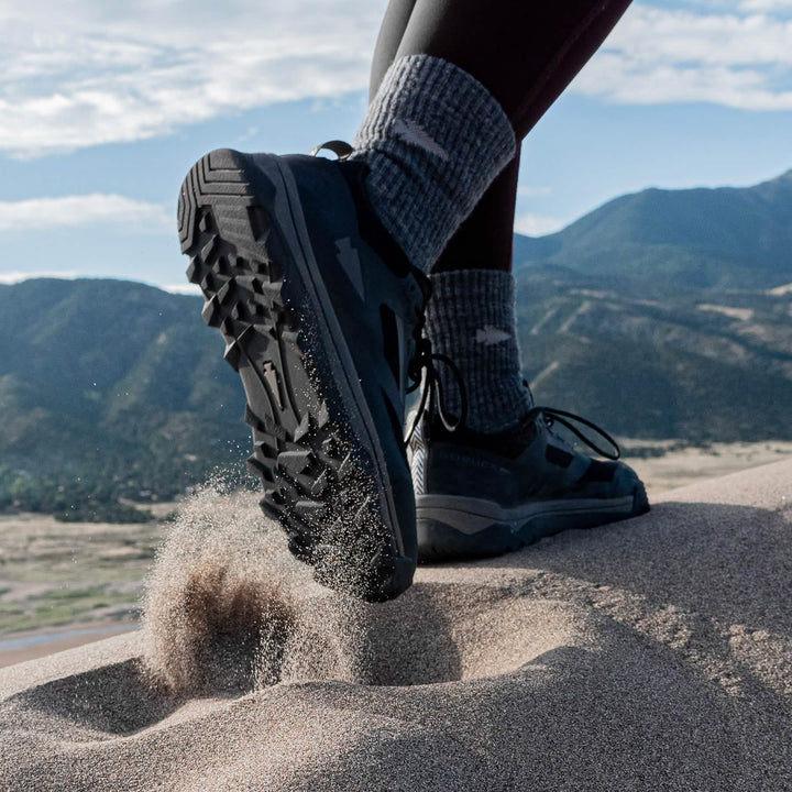 A person hiking on sand dunes in GORUCK Mackall - Black + Forged Iron trail shoes, showcasing the triple compound outsole as sand is kicked up, with majestic mountains in the background.