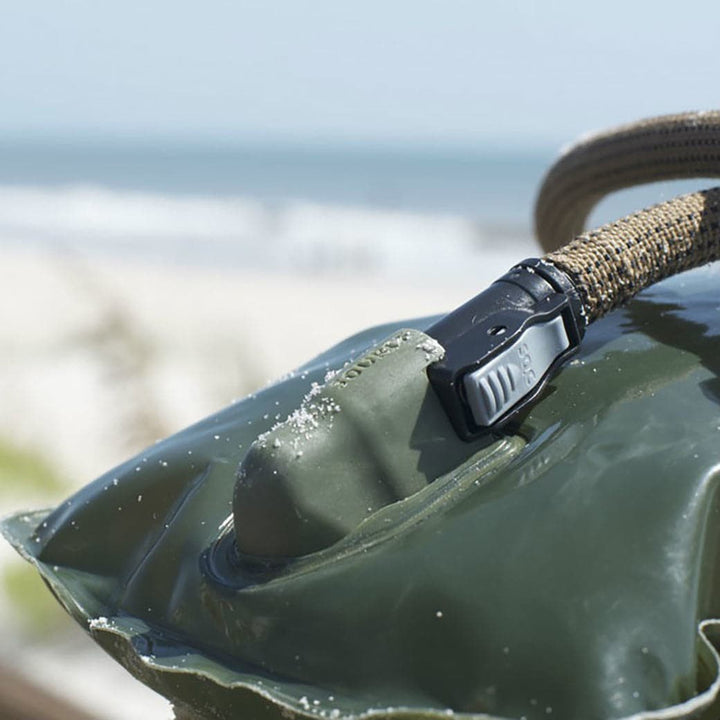 Close-up of a Source Hydration Bladder in green with a hose, showcasing antimicrobial technology, on a sandy beach with the ocean in the background.