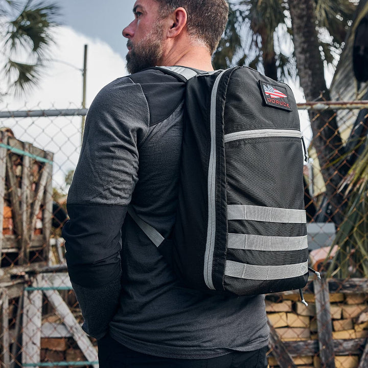 A man carrying a GORUCK Bullet Ruck Laptop backpack in Ripstop Nylon - 16L, designed with lumbar support, stands by a chain-link fence with palm trees in the background.