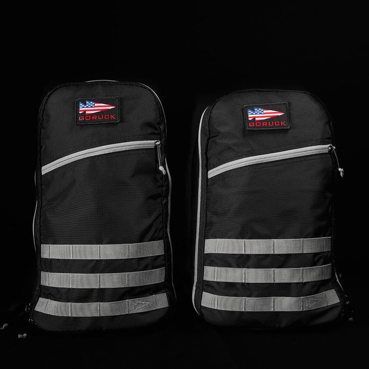 Displayed against a dark background, two identical GORUCK Bullet Ruck Laptop backpacks, crafted from durable 16L Ripstop ROBIC® Nylon with gray straps and zippers, stand out. Each backpack features a USA flag patch alongside the GORUCK label. With their rugged, minimalist design, they offer lumbar support and a laptop compartment for maximum utility.