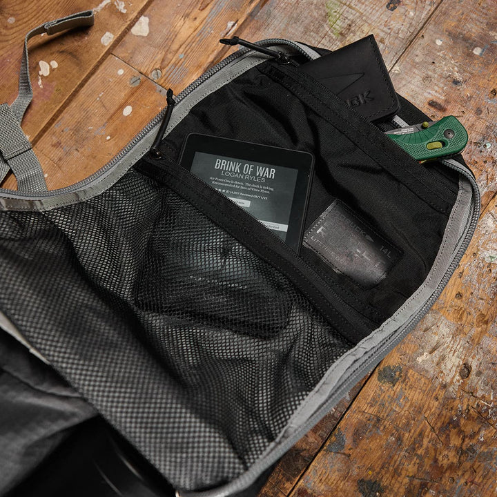 On a wooden floor, the GORUCK Bullet Ruck Laptop backpack in Ripstop Nylon (16L) is opened to showcase its water-resistant exterior. Inside, a Kindle, knife, and wallet are neatly organized in mesh pockets, along with an additional compartment for your laptop and tech essentials.