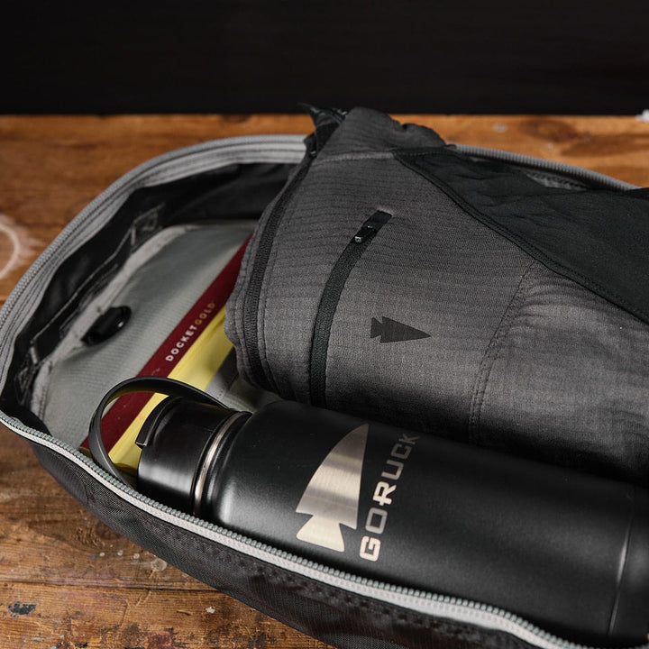 On a wooden surface, the Bullet Ruck Laptop - Ripstop Nylon - 16L by GORUCK is open, revealing a book and a black GORUCK water bottle inside, complete with a laptop compartment and designed to provide lumbar support.