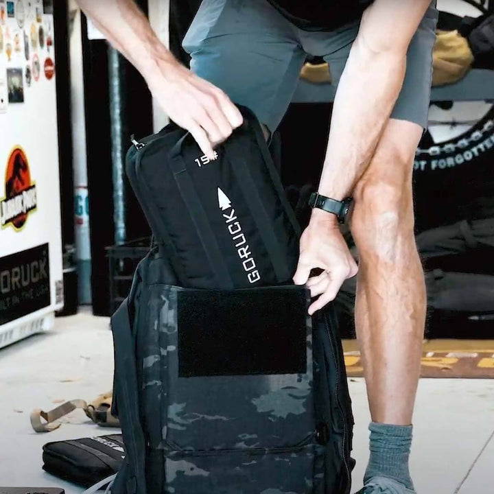 Indoors, a person is carefully placing a GORUCK bag into a larger camo backpack, ensuring there's enough room for the Sand Ruck Plates, which will make the rucking experience challenging yet rewarding.