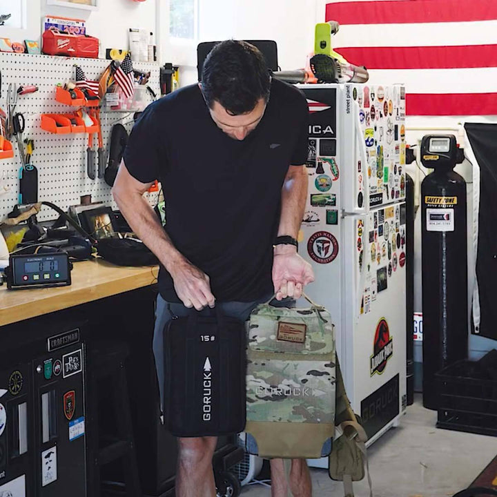 A man stands in a workshop, holding two GORUCK bags equipped with a portable Sand Ruck Plate. Behind him, the American flag waves proudly amidst an array of eclectic stickers.