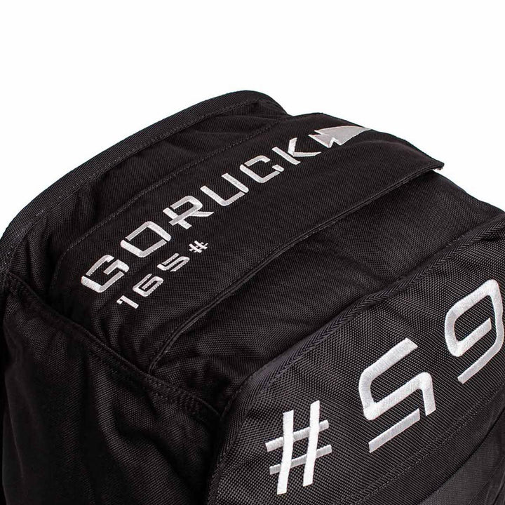 A close-up of a black Sand Tombstones by GORUCK showcases white embroidered text, including GORUCK, 165, and partial text 165 on the fabric. Its rugged design features a multilayer closure system, making it perfect for loaded carries and inspired by the durability of the Husafell Sandbag.