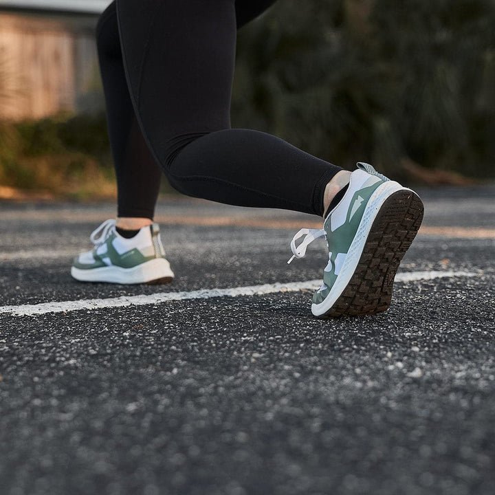 A person wearing black leggings and green Men's Rough Runner - Seaspray sneakers by GORUCK, equipped with Gradient Density EVA soles, is running on a paved road. The angle focuses on their legs and shoes, capturing the motion of this Rough Runner. White lines mark the road surface.