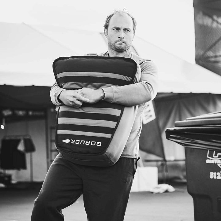 A man is standing outdoors, gripping a large protective pad labeled GORUCK in black and white. With a focus akin to tackling Sand Tombstones, he stands ready, as if for loaded carries. Behind him are a tent and a large bin, adding to the scene's rugged aura.