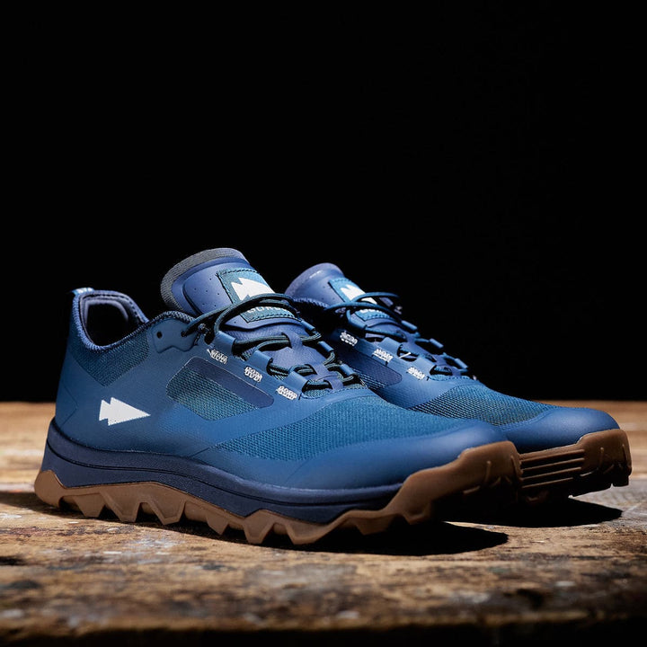 The Mackall trail running shoes by GORUCK in Legion Blue + Gum rest on a wooden surface against a dark background, their robust brown soles revealing the all-terrain challenges they are designed to overcome.