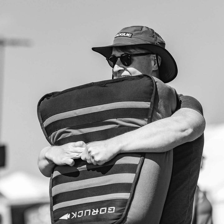 A person wearing sunglasses and a hat is holding a GORUCK Sand Tombstone, reminiscent of a Husafell Sandbag. The black-and-white image's blurry background hints at an outdoor setting focused on loaded carries.