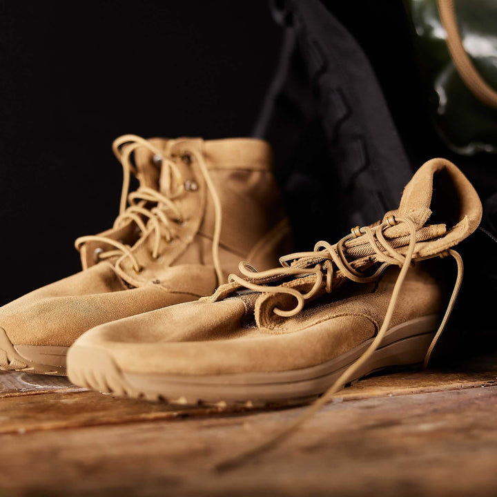 The GORUCK MACV-1 Hi-Speed Mid Top tan suede boots with laces rest elegantly on wood, complemented by a dark bag in the background. Featuring an EVA midsole, these lightweight boots offer comfort and style.