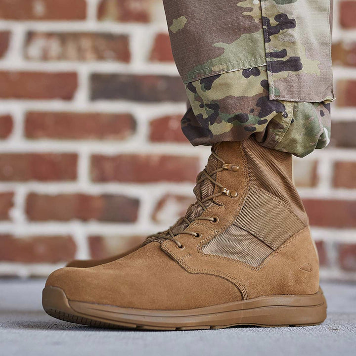 A close-up of a person wearing brown MACV-1 Hi-Speed - High Top rucking boots by GORUCK and camouflage pants against a brick wall showcases their AR670-1 compliance while maintaining a lightweight feel.