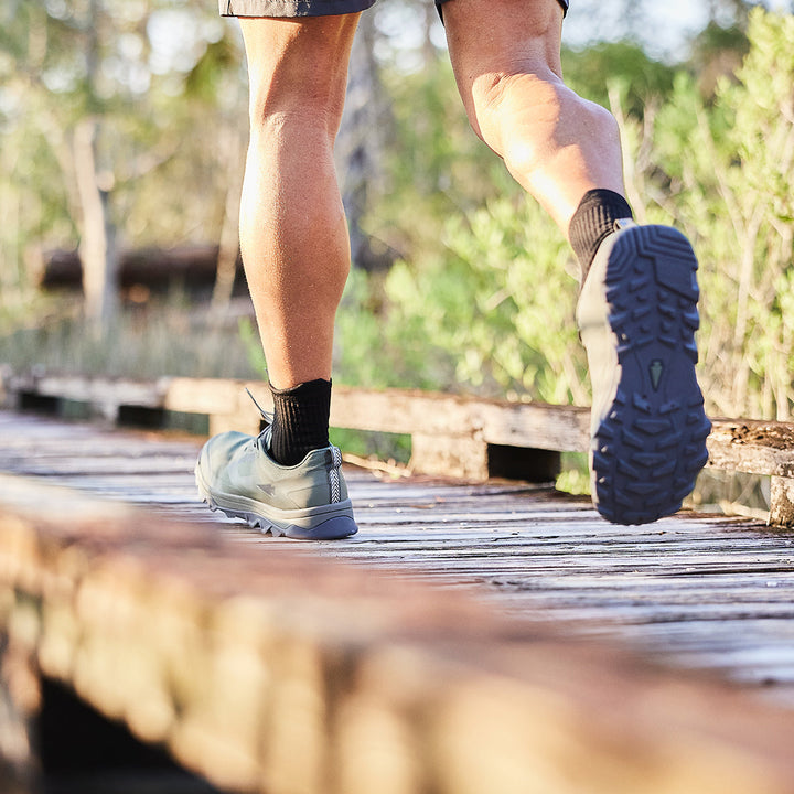 A person strides along a wooden path in the woods, wearing GORUCK Mackall shoes in Earth and Forged Iron colors with black socks, reminiscent of Special Forces training sessions.