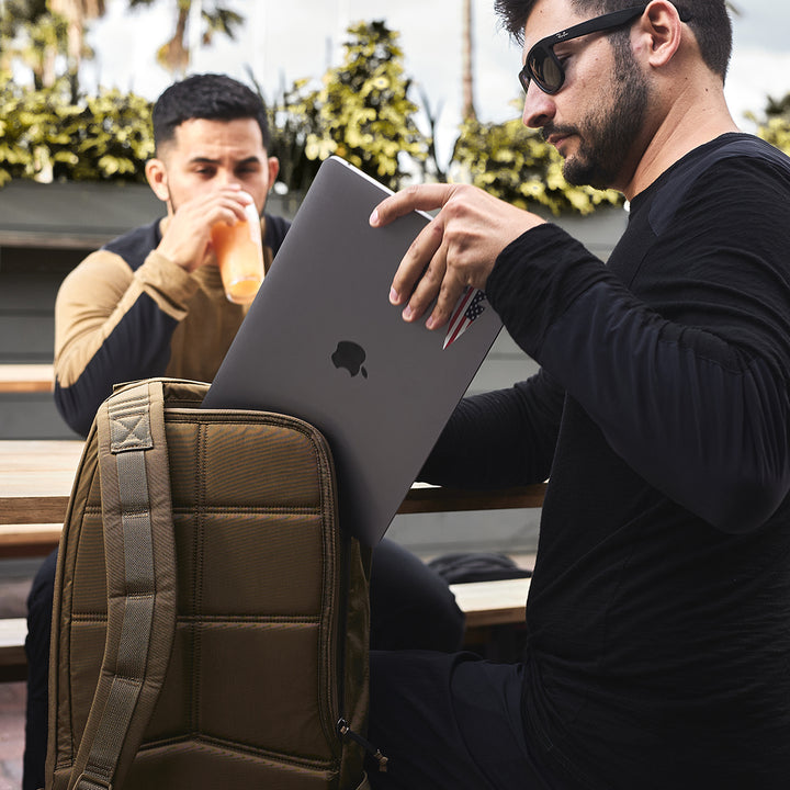 Two men sit outdoors, one wearing sunglasses uses a laptop tucked safely in a GORUCK Bullet Ruck Laptop - 16L with a bombproof compartment, the other sips from a cup, enjoying the day. Lush plants create a serene backdrop to their relaxed yet productive setting.