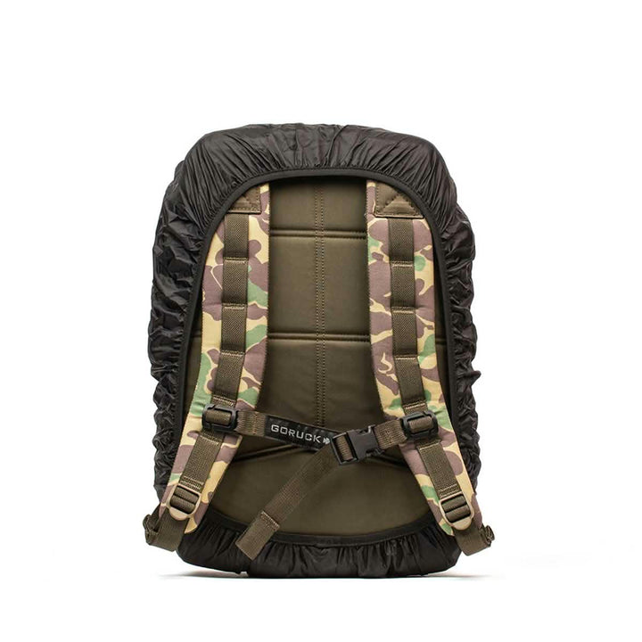 A backpack featuring a camouflage pattern is accompanied by the GORUCK's Ruck Rain Cover, a lightweight black waterproof cover. The rear of the backpack includes padded straps and a waist buckle for added support. The camouflage design integrates shades of green, tan, and brown to create a rugged outdoor appearance.