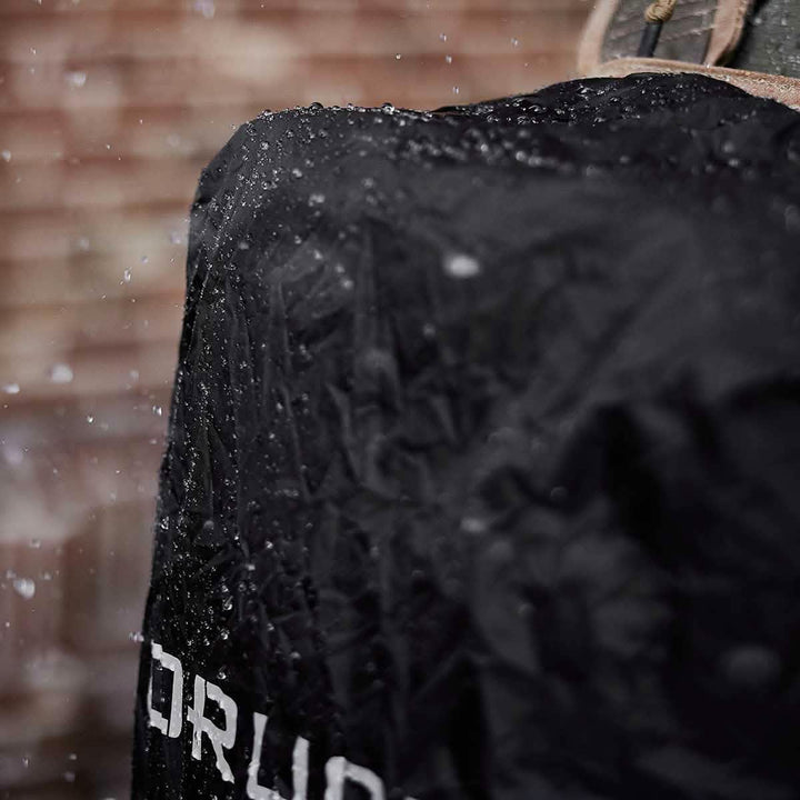 A close-up of the Ruck Rain Cover by GORUCK, a black waterproof cover, glistens with droplets. Text from the partially visible logo stands out against the blurred backdrop of a brick wall, as raindrops cascade down, showcasing its lightweight design.