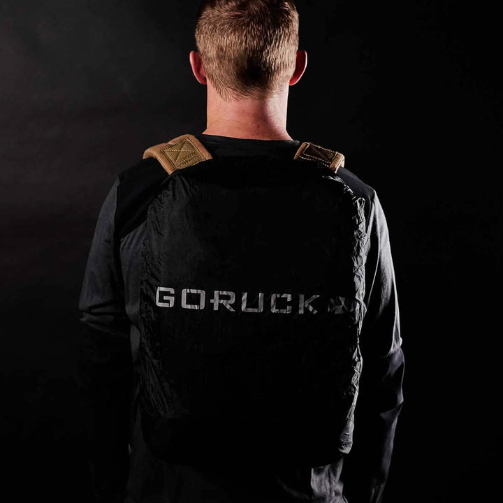 A person with short hair, seen from behind, is sporting a black GORUCK backpack. The image's dark background emphasizes the rucksack's reflective logo and tan shoulder straps, while suggesting that a lightweight Ruck Rain Cover from GORUCK keeps it protected.