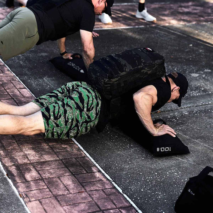 A person doing push-ups with a heavy GORUCK backpack, enhanced by Simple Training Sandbags, outdoors on a paved path.