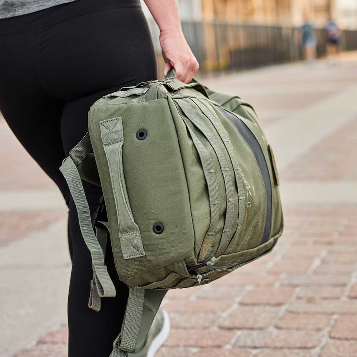 A person walks holding a green Rucker 4.0 backpack by GORUCK, paired with black leggings and white sneakers, ready for a day of rucking.