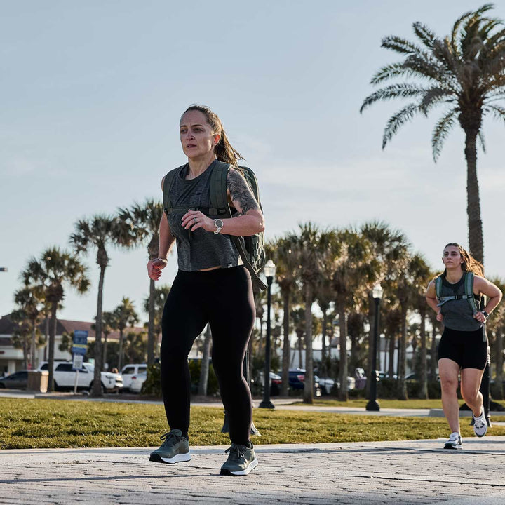 Two people jog on a paved park path, palm trees swaying gently, effortlessly incorporating GORUCK Rucker 4.0 into their workout for added intensity.