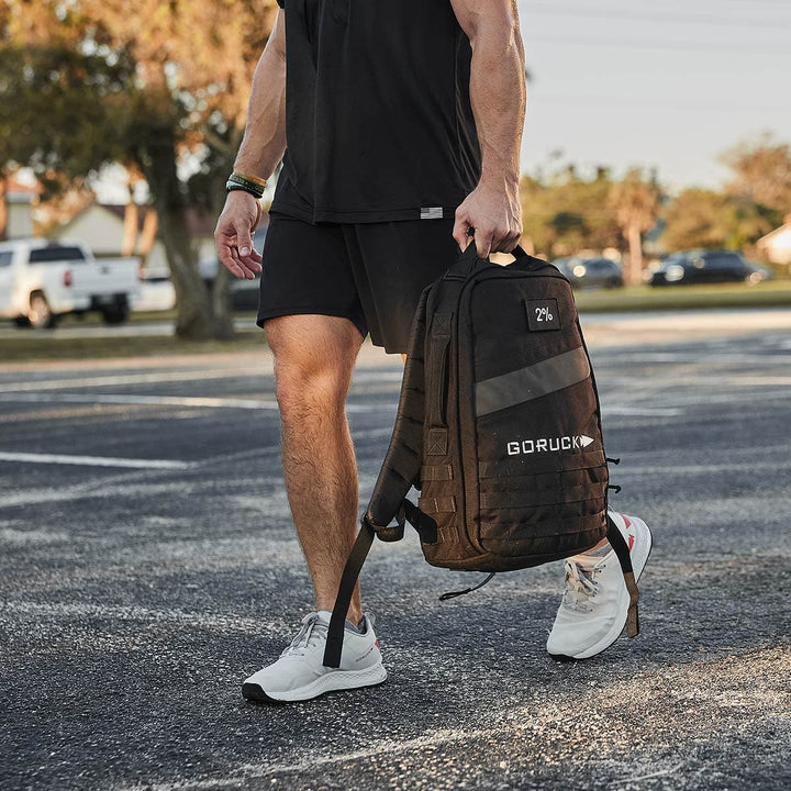 A person wearing black shorts and a T-shirt walks across a parking lot holding a black Rucker 4.0 backpack from GORUCK. They are sporting white sneakers with black and red accents, ideal for rucking. In the background, cars and trees are visible under the clear sky.