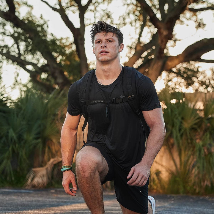 Dressed in a black workout outfit and equipped with a Rucker 4.0 from GORUCK, a person is exercising outdoors in a park setting, performing lunges with their backpack. As the sun sets, it casts a warm glow over the determined individual, enhancing the serene yet invigorating ambiance of the moment.