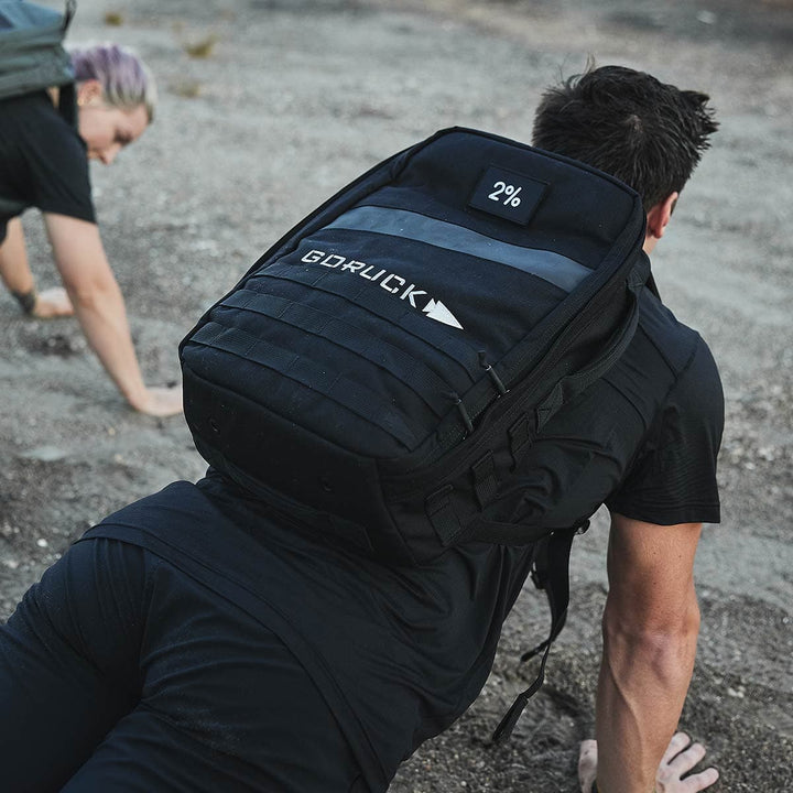 Two people are exercising outdoors on a dirt path. Leading the way, the person wearing a black shirt and carrying a GORUCK Rucker 4.0 demonstrates their commitment to rucking. In the background, adding depth to the image, is another individual with their hair tied back, slightly out of focus.