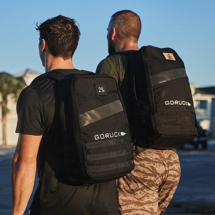 Two men walking outdoors, each sporting a black GORUCK Rucker 4.0 backpack. One wears a black shirt while the other dons a green shirt with camo pants. They stride through a sunlit, urban setting with buildings in the background, fully embracing the spirit of rucking.