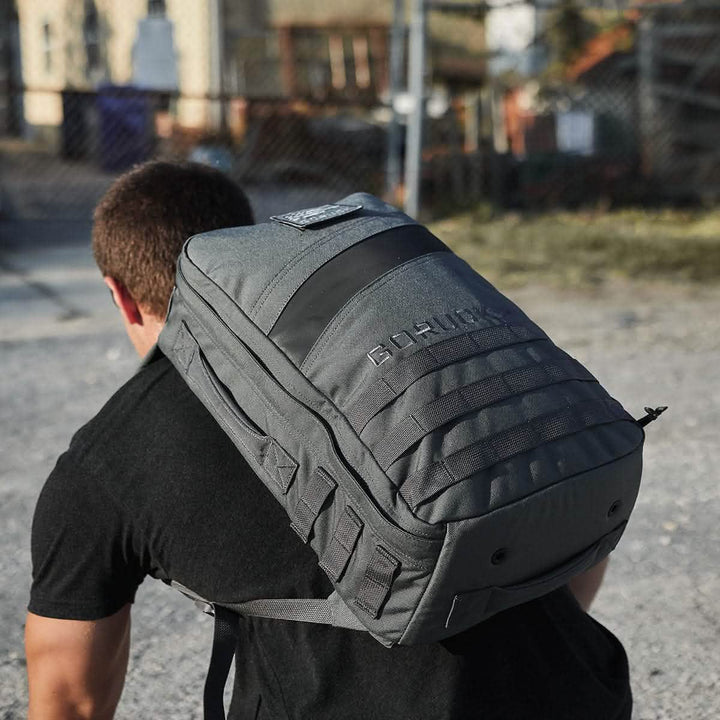 A person in a black shirt is carrying the large Rucker 4.0 by GORUCK on their back. They're outdoors on a gravel surface, with a blurred fence and building in the background.