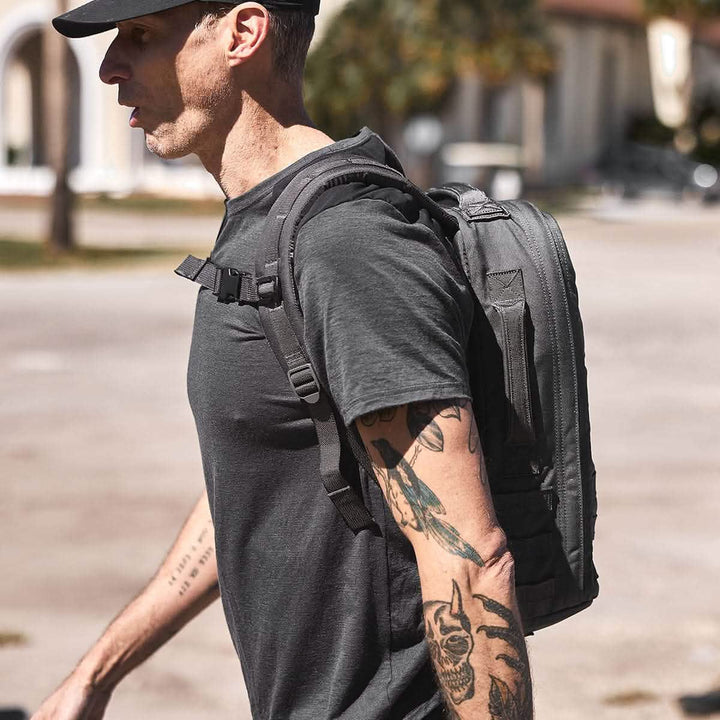 A person wearing a black cap and a black T-shirt is walking outdoors with a Rucker 4.0 backpack by GORUCK. His arms are covered in tattoos, and the background is slightly blurred with trees and buildings.