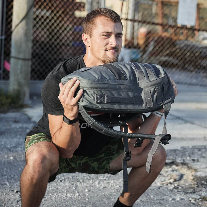 A person is performing a squat exercise outdoors, holding a gray GORUCK Rucker 4.0 backpack close to their chest. They are wearing a black shirt and green camouflage shorts, with a chain-link fence and some industrial background elements visible.