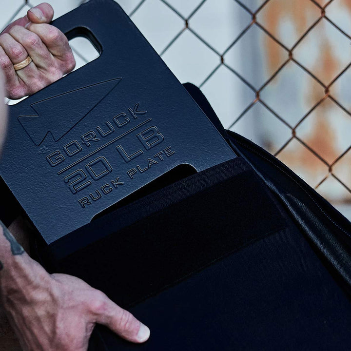 A rucker carefully places a 20 lb Ruck Plates - LB from GORUCK into their bag by a chain-link fence.