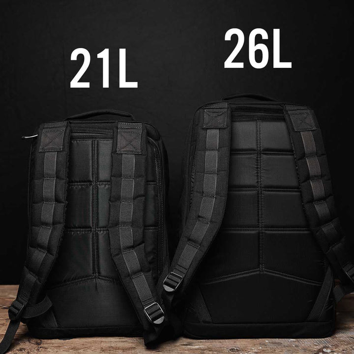 Two black GR1 USA - Cordura (The Original Ruck) backpacks from GORUCK are displayed against a dark background. The left backpack, ideal for a GORUCK Challenge, is labeled 21L, while the right one is labeled 26L GR1, highlighting their different sizes. Both Special Forces-inspired backpacks feature padded straps and are placed on a wooden surface.