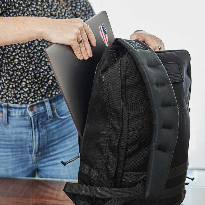 An individual is meticulously placing a laptop into a sleek black GR1 USA rucksack by GORUCK on the table, highlighting the durability of its 210D CORDURA fabric.