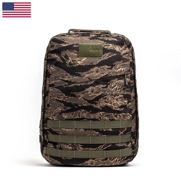 The GR1 USA by GORUCK showcases a camo-patterned rucksack crafted with robust 210D CORDURA fabric and accented with green straps against a white background. A small U.S. flag on the top left corner lends a patriotic touch to its durable design.