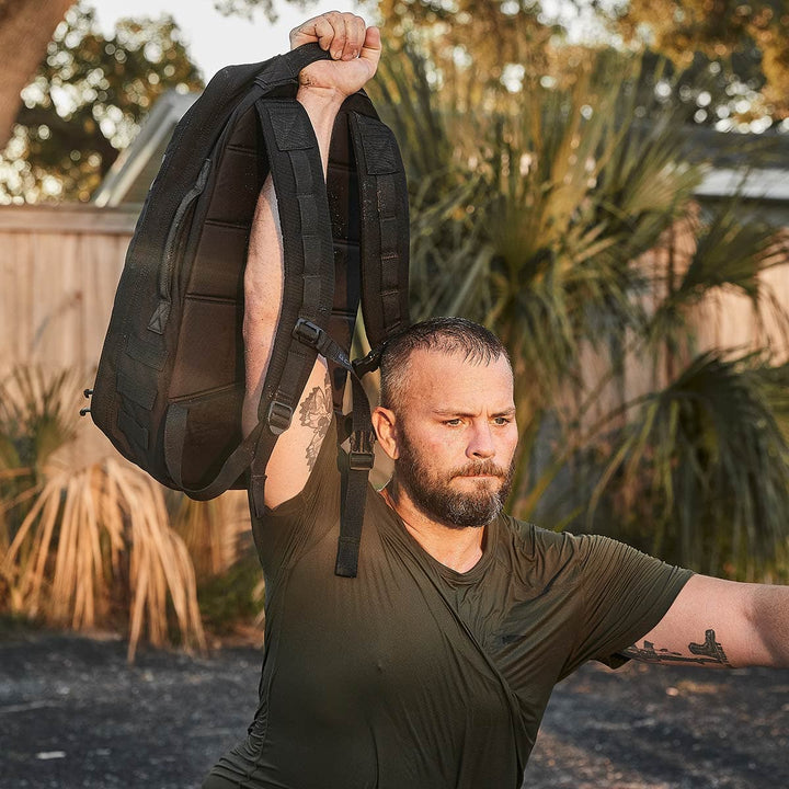 Outdoors, a bearded man triumphantly lifts his GORUCK Rucker 4.0 rucksack overhead with one hand. His dark, wet shirt suggests the exertion from rucking, and tattoos cover his arm. The background is filled with greenery and a wooden fence, resembling a yard or park setting.