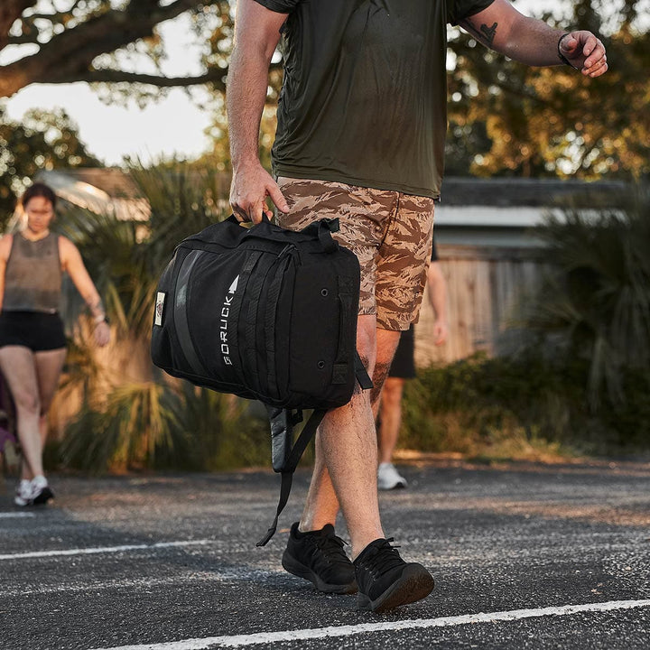 A person carrying a GORUCK Rucker 4.0 backpack, dressed in camouflage shorts and black shoes, strolling outside on a sunny day—ideal for a rucking adventure.