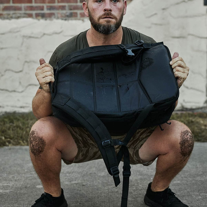 A man squats outdoors with the GORUCK Rucker 4.0 slung over his shoulders, dressed in a green T-shirt and camo shorts, ready for a day of intense rucking.