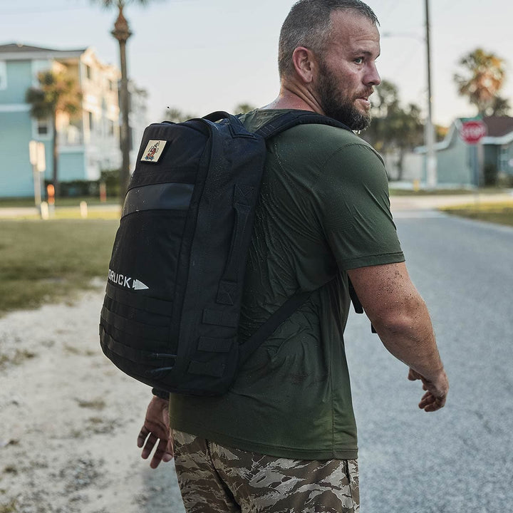 A man with a short beard is dressed in a wet green T-shirt and camo shorts, carrying his black GORUCK Rucker 4.0 backpack as he walks down the street. In the background, houses, palm trees, and a stop sign are visible under a clear sky.