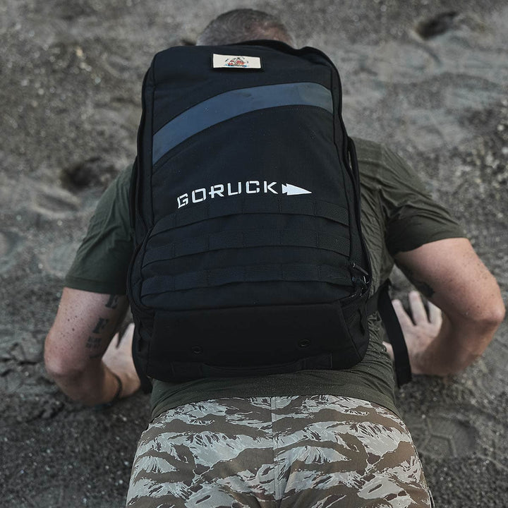 A person wearing camouflage pants and a Rucker 4.0 backpack is performing push-ups on a sandy surface, capturing the essence of rucking. The black backpack proudly displays the GORUCK brand name.