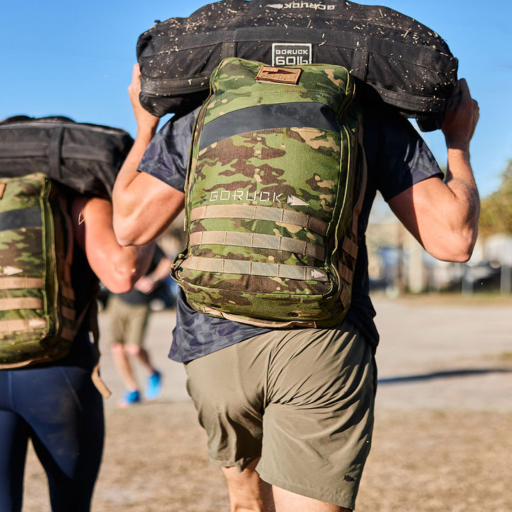 Two individuals, each equipped with GORUCK Rucker 4.0 backpacks, carry heavy sandbags as they engage in a challenging outdoor rucking event.