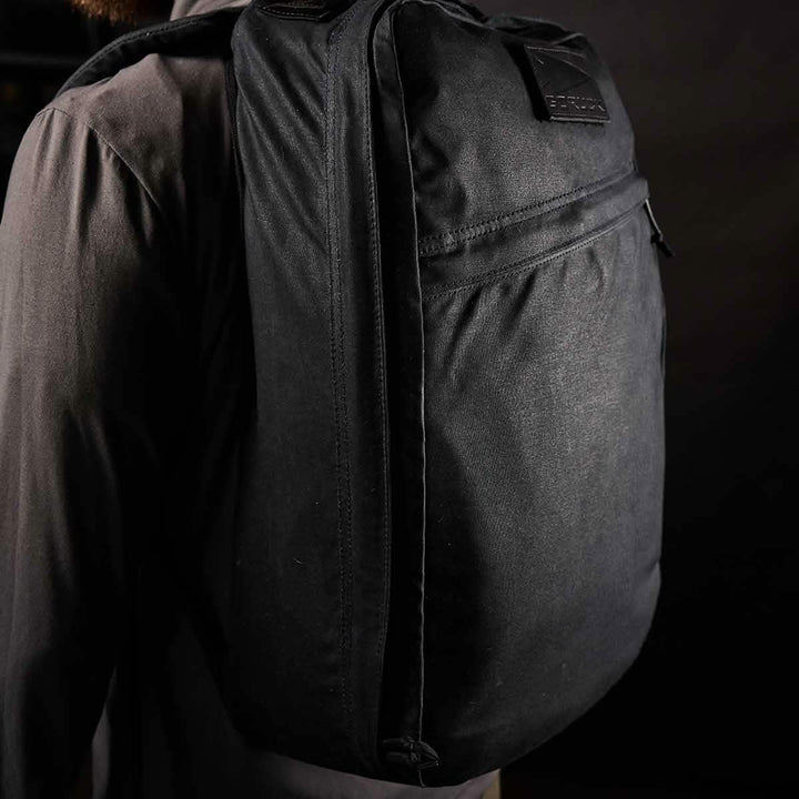 A person stands against a black background, wearing GORUCK's GR1 USA - Heritage dark jacket with a heritage vibe, perfectly paired with a black waxed canvas rucksack.