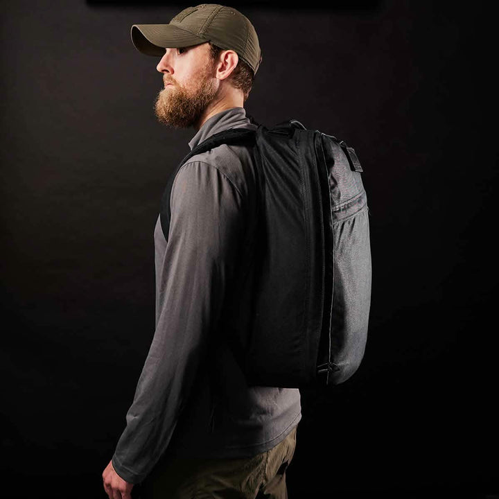 A bearded man in a cap and jacket carries the GORUCK GR1 USA - Heritage rucksack made from rugged Waxed Duck Canvas, set against a dark background.