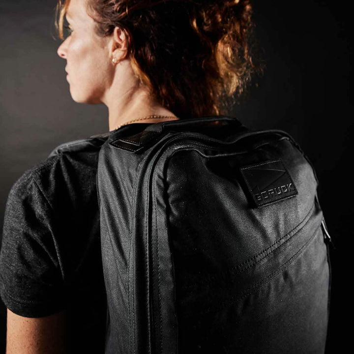 A person with curly hair stands sideways against a dark background, wearing a GR1 USA - Heritage rucksack from GORUCK.