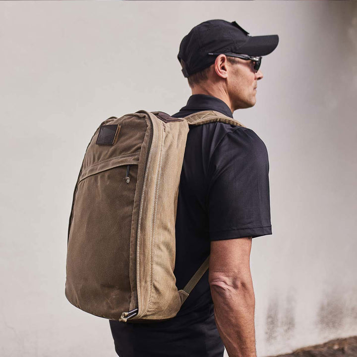 A man in a black cap and shirt, wearing sunglasses, is carrying a large beige GR1 USA - Heritage rucksack from GORUCK against a plain background.