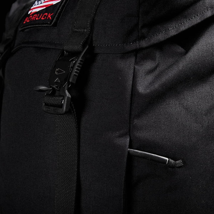 Close-up image of the GORUCK M23 - Cordura rucksack showcasing a strap with a buckle and a red patch bearing white letters, partially visible. The rugged fabric suggests the use of durable materials, ensuring a resilient design.