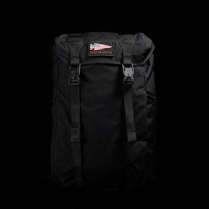 The M23 - Cordura by GORUCK is a modern black rucksack featuring dual straps and buckle closures. It showcases a patch with an American flag and the GORUCK logo on the front. Crafted from durable materials, this rucksack is designed for both style and endurance, set against a black background.