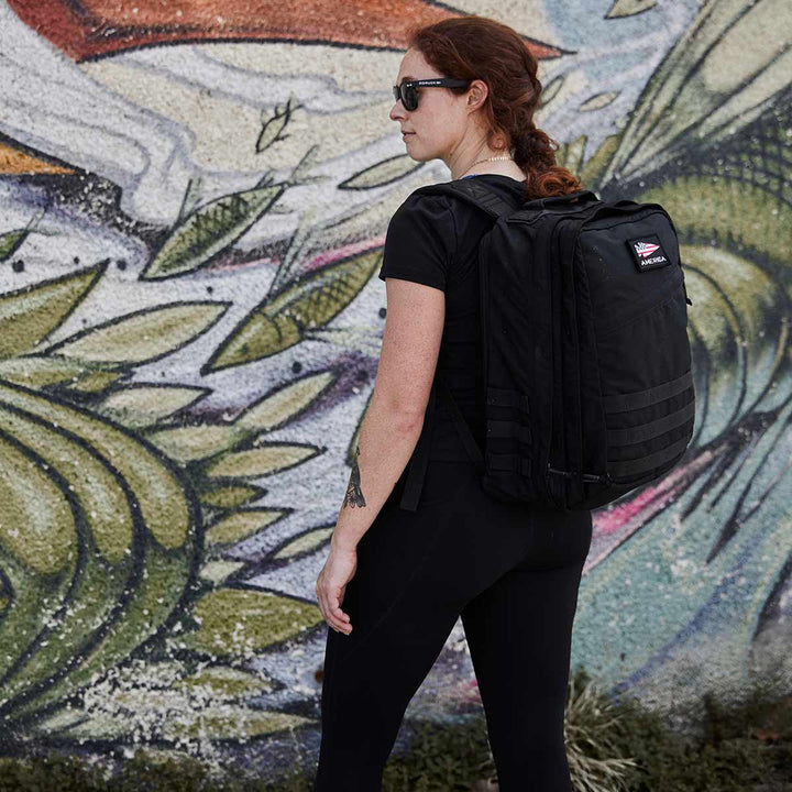 A woman with a black backpack stands in front of a colorful graffiti art wall, her GORUCK GR2 - USA's 210D CORDURA® design perfectly blending function with urban flair.