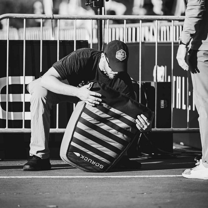 A person crouches on the ground, gripping a Sand Tombstones backpack by GORUCK with a multilayer closure system and striped patterns, next to a metal barricade. The scene unfolds outdoors, evoking an old-world charm in black and white.