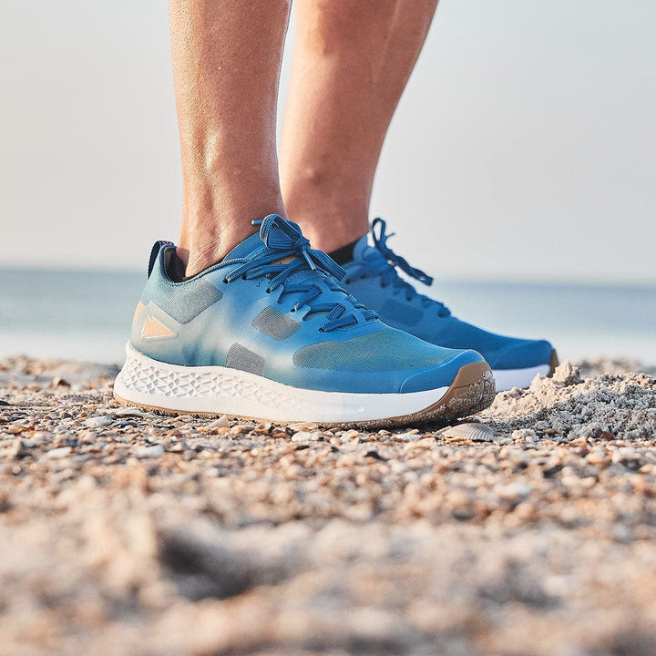 A person stands on a sandy beach wearing GORUCK's Men's Rough Runner in Tidal Blue. These athletic shoes boast a textured white sole with Gradient Density EVA, ideal for long-distance running. Small pebbles scatter the sand, complementing the peaceful backdrop of the ocean and sky.